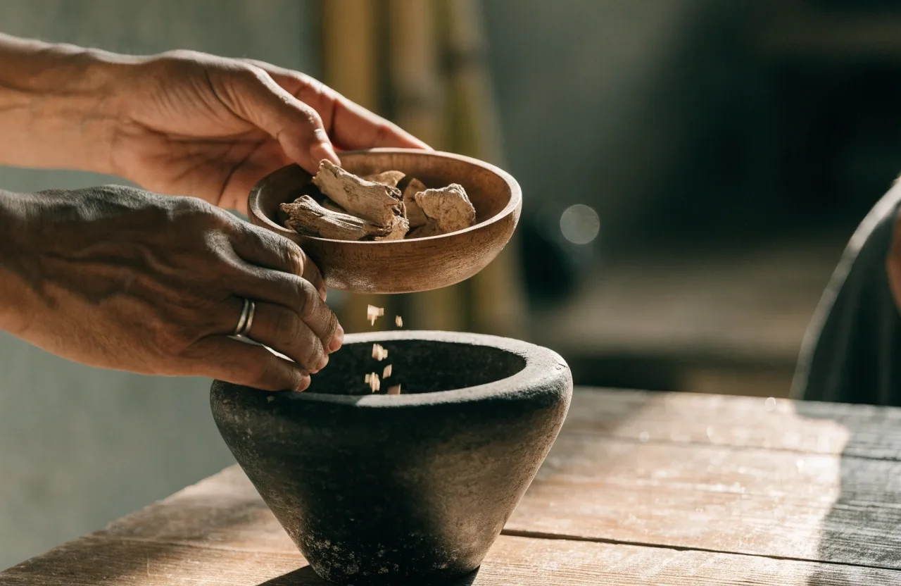 Fine milled botanical powder pouring from a wooden scoop into a small dark ceramic bowl in warm light